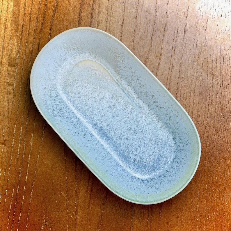 May include: An oval-shaped ceramic serving dish with a light blue and white speckled glaze. The dish has a slightly raised rim and a recessed center. The dish is approximately 10 inches long and 5 inches wide.