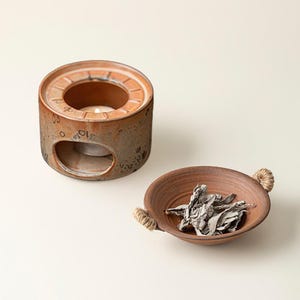 May include: A ceramic essential oil diffuser with a tea light candle inside, next to a small brown bowl with rope handles, filled with dried sage leaves. The diffuser and bowl are brown with a textured finish.