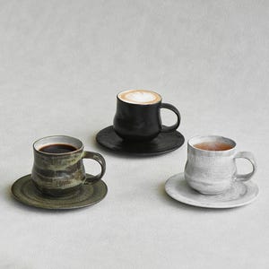以下が含まれることがあります： 黒いハンドルと「But First, Coffee」のテキストが黒い太字で書かれた白いセラミックマグ。マグカップは木製の表面に置かれています。