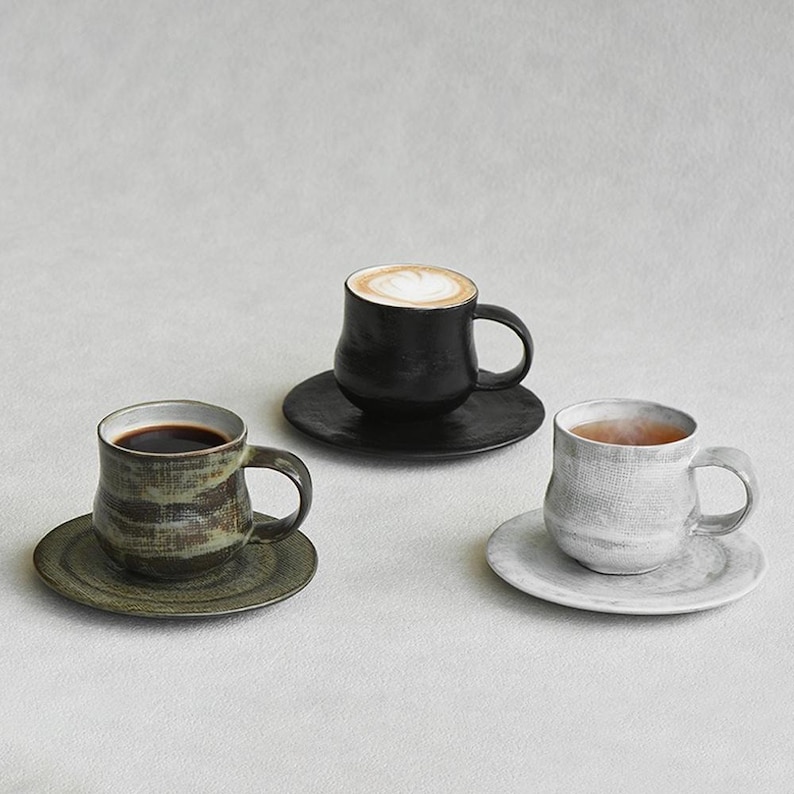 K&ouml;nnte beinhalten: Eine wei&szlig;e Keramik-Tasse mit schwarzem Henkel und dem Text "But First, Coffee" in schwarzer, fetter Schrift. Die Tasse steht auf einer Holzoberfl&auml;che.
