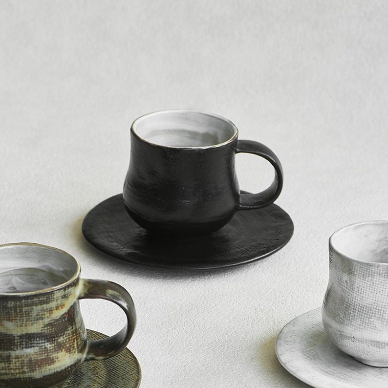 K&ouml;nnte beinhalten: Ein Set handgefertigter Keramik-Kaffeetassen und Untertassen. Die schwarzen Tassen haben wei&szlig;e Innenr&auml;ume und stehen auf passenden schwarzen Untertassen. Weitere Tassen sind in Braun- und Wei&szlig;t&ouml;nen gehalten. Die Tassen haben ein rustikales, handwerkliches Aussehen.