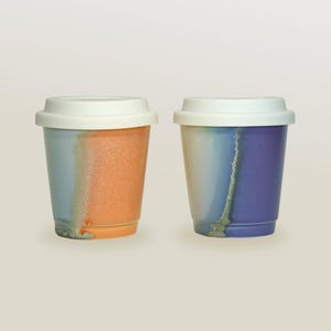 May include: Two ceramic coffee mugs with white lids. The mug on the left has a blue and orange glaze. The mug on the right has a blue, purple, and white glaze.