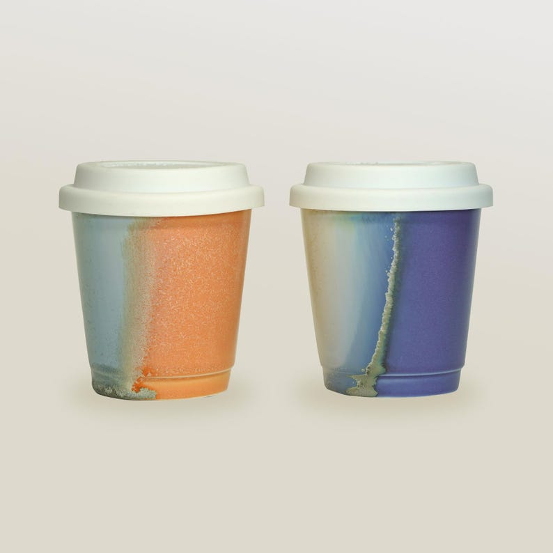 May include: Two ceramic coffee mugs with white lids. The mug on the left has a blue and orange glaze. The mug on the right has a blue, purple, and white glaze.