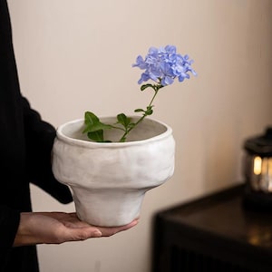 Puede incluir: Un jarrón de cerámica blanca con una superficie irregular y texturizada. El jarrón tiene forma de un tazón ancho con una base estrecha. Un solo tallo de flores azul claro está dispuesto en el jarrón.