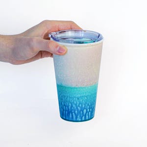 May include: A tall, reusable tumbler with a clear lid is held by a hand. The tumbler features a gradient design, transitioning from white to turquoise to a textured blue base. The tumbler is designed for cold beverages.