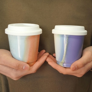 May include: Two ceramic reusable coffee cups with white lids. The cup on the left has a blue, white, and orange glaze. The cup on the right has a blue, white, and purple glaze.