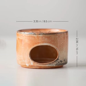 May include: A cylindrical ceramic container with an oval opening. The container is orange with dark brown accents and measures 3.35 inches wide and 2.36 inches tall. The handmade pottery has a rustic, textured appearance.