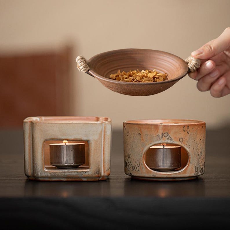 May include: Two ceramic oil warmers with lit tea lights. One is square, the other is round. A brown ceramic bowl with rope handles and dried herbs is held above the warmers. The warmers are beige and brown.