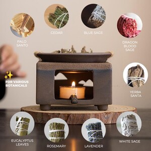 Op de afbeelding: Een vierkante, donkerbruine keramische etherische olie diffuser met een brandende kaars, omringd door verschillende botanische producten. De botanische producten omvatten Palo Santo, Ceder, Blauwe Salie, Drakenbloed Salie, Yerba Santa, Eucalyptusbladeren, Rozemarijn, Lavendel en Witte Salie.