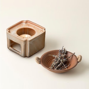 May include: A hexagonal ceramic essential oil diffuser with a lit candle inside, next to a small brown bowl with rope handles and dried lavender. The diffuser and bowl are in shades of brown and tan.