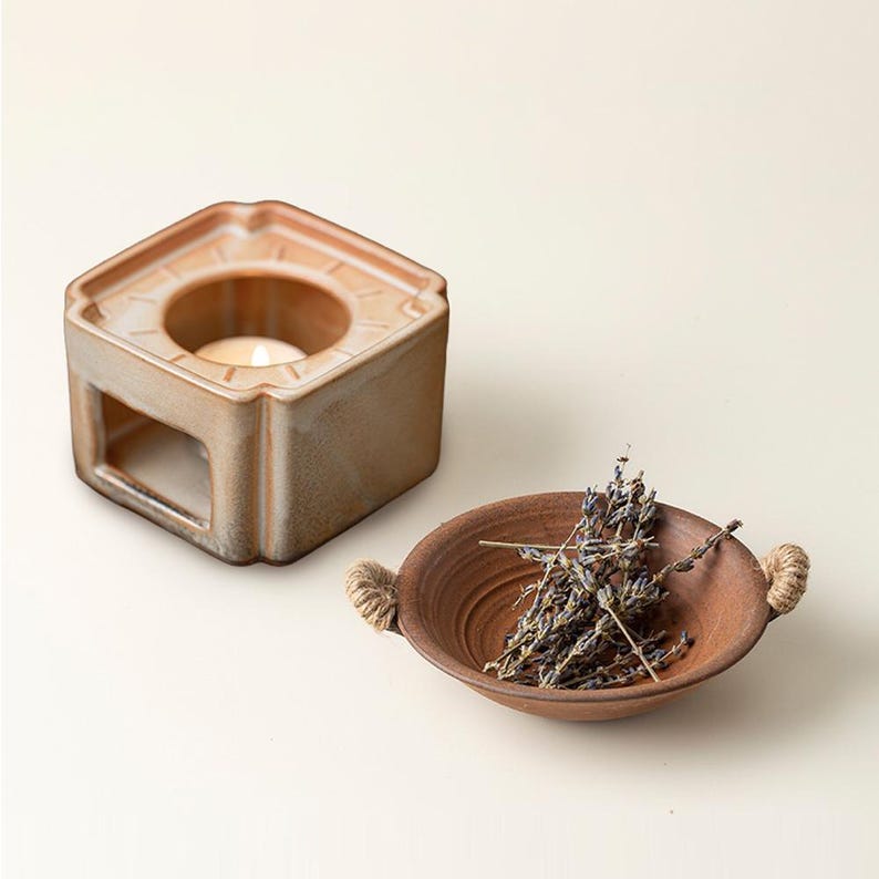 May include: A hexagonal ceramic essential oil diffuser with a lit candle inside, next to a small brown bowl with rope handles and dried lavender. The diffuser and bowl are in shades of brown and tan.