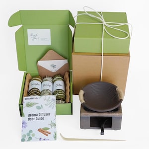 Op de afbeelding: Een open groene doos onthult eucalyptus, blauwe salie en cederbundels, samen met een aromadiffuser en een gebruikershandleiding. Een bruine envelop en kaart zijn ook zichtbaar. Twee groene dozen zijn vastgebonden met wit touw.
