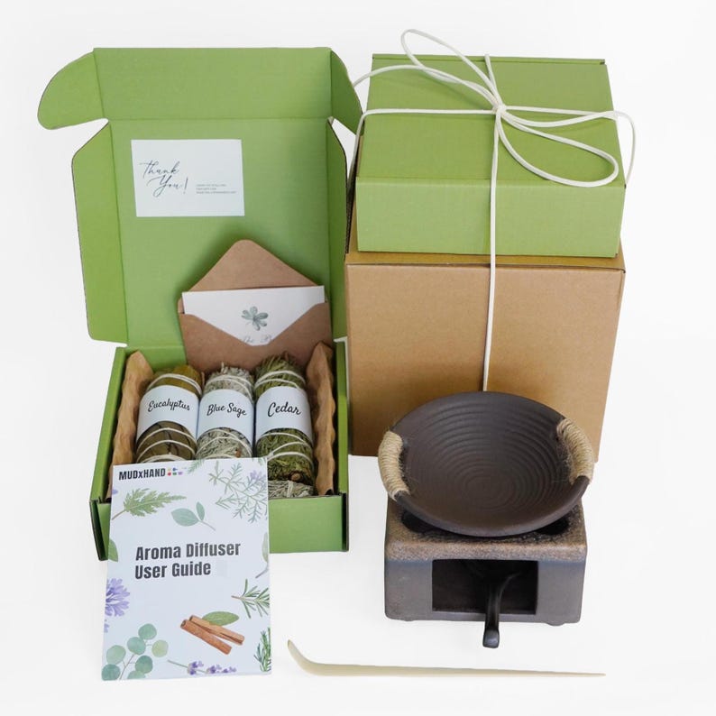 Op de afbeelding: Een open groene doos onthult eucalyptus, blauwe salie en cederbundels, samen met een aromadiffuser en een gebruikershandleiding. Een bruine envelop en kaart zijn ook zichtbaar. Twee groene dozen zijn vastgebonden met wit touw.