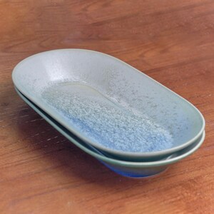 May include: Two oval-shaped ceramic serving dishes. The dishes are stacked, showcasing a gradient of colors from light blue to a deeper blue. The surface has a speckled texture, adding visual interest to the tableware.