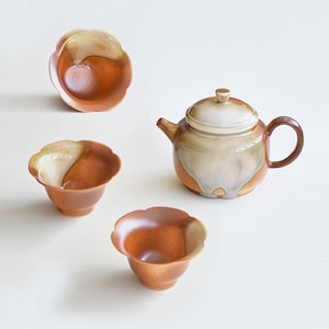 Puede incluir: Una tetera de cerámica marrón y blanca con tapa y un juego a juego de tres tazas de té pequeñas. Las tazas tienen un borde festoneado y están diseñadas para parecerse a una flor.