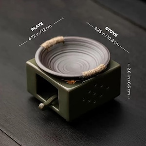 Op de afbeelding: Olijfgroene keramische kachel met een donkergrijze plaat, met een diameter van 12 cm. De plaat is versierd met touw en staat op een rechthoekige basis. De kachel is 10,8 cm breed en 6,6 cm hoog.