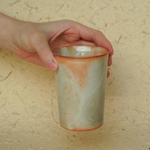 Może przedstawiać: Ceramiczny kubek z jasnozieloną glazurą i czerwono-pomarańczową krawędzią. Kubek jest lekko przezroczysty i ma marmurkowy wygląd.
