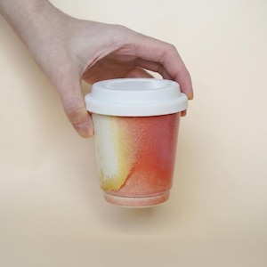 May include: A ceramic travel mug with a white lid. The mug features a gradient design with colors ranging from white to yellow and red. The mug is held by a hand against a neutral background.