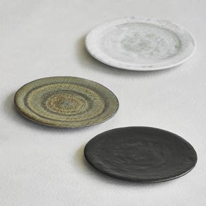K&ouml;nnte beinhalten: Drei Keramikplatten in einer neutralen Farbpalette. Die Platten sind rund mit einer strukturierten Oberfl&auml;che. Eine Platte ist wei&szlig; mit einem gr&uuml;nen Zentrum, eine weitere olivgr&uuml;n und die dritte schwarz.