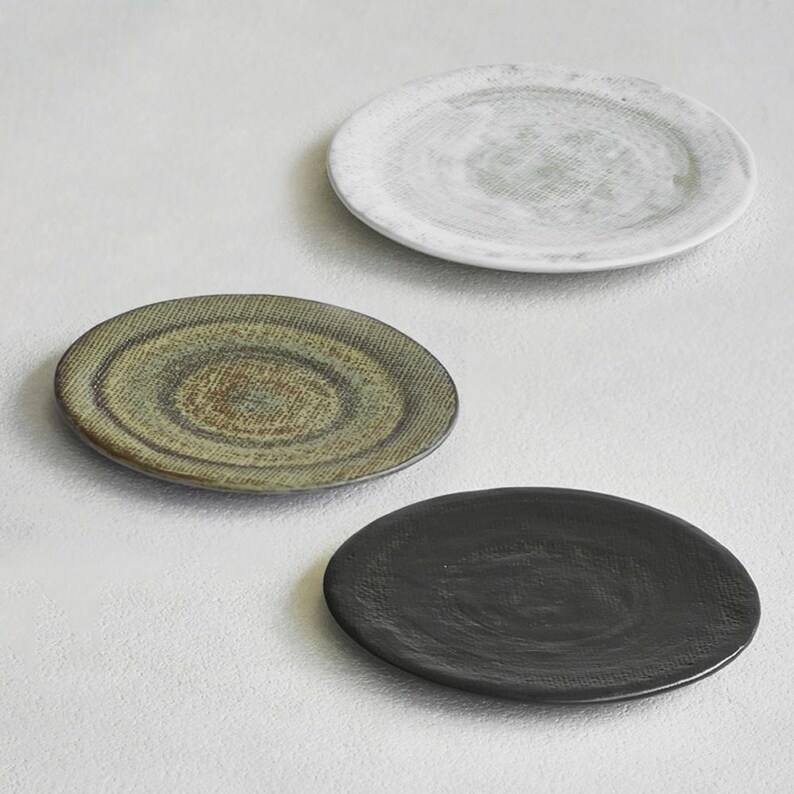 K&ouml;nnte beinhalten: Drei Keramikplatten in einer neutralen Farbpalette. Die Platten sind rund mit einer strukturierten Oberfl&auml;che. Eine Platte ist wei&szlig; mit einem gr&uuml;nen Zentrum, eine weitere olivgr&uuml;n und die dritte schwarz.
