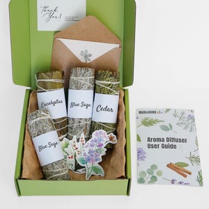 Op de afbeelding: Een groene doos met bundels eucalyptus, blauwe salie en ceder, elk gelabeld met zijn naam. De doos bevat ook stickers, een kleine envelop en een gebruikershandleiding voor een aromadiffuser. De doos is open en onthult de inhoud.