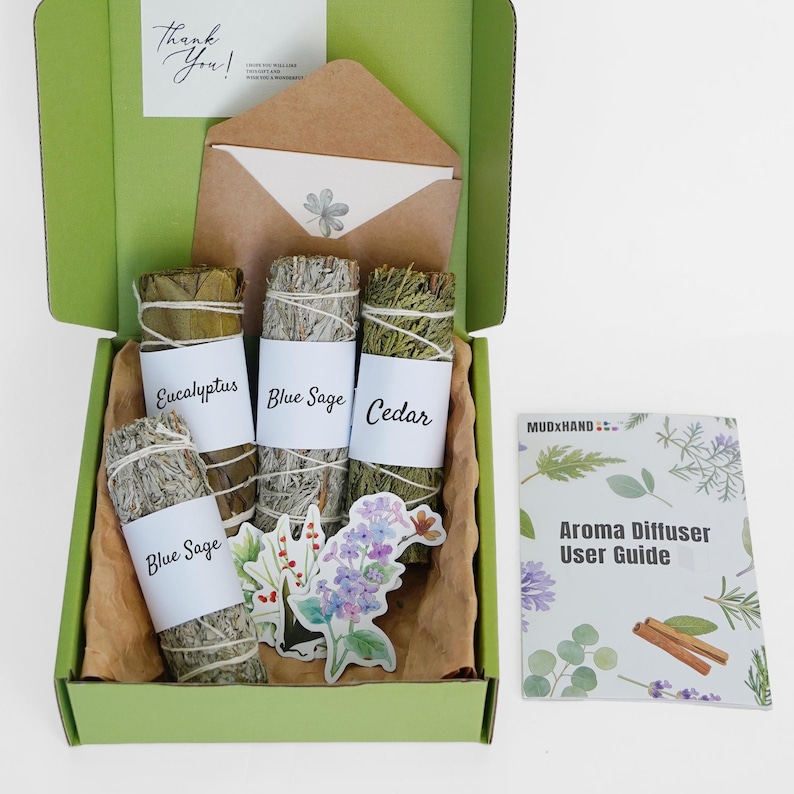 Op de afbeelding: Een groene doos met bundels eucalyptus, blauwe salie en ceder, elk gelabeld met zijn naam. De doos bevat ook stickers, een kleine envelop en een gebruikershandleiding voor een aromadiffuser. De doos is open en onthult de inhoud.