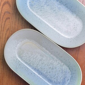 May include: Two oval-shaped serving platters with a speckled, light blue and white glaze. The platters have a slightly raised rim and a smooth, elongated interior. The background is a warm, wooden surface.