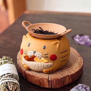 Peut inclure: Un brûle-parfum en céramique jaune en forme de chat tenant un poisson, avec une bougie chauffe-plat allumée à l'intérieur. Un plat en céramique beige, rond et peu profond, doté de deux petites anses, est posé au-dessus avec des herbes.