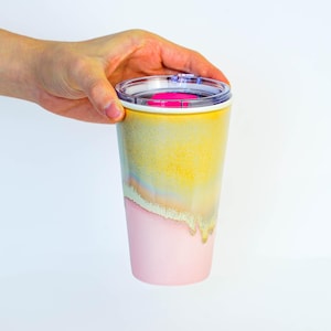 May include: A colorful travel mug with a clear lid and a pink accent. The mug has a gradient design, transitioning from yellow to a light pink. The mug is held by a hand against a white background.
