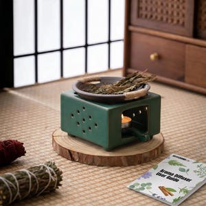 May include: A square, dark green ceramic aroma diffuser with perforated sides and a central opening containing a lit tea light candle. A shallow, dark brown ceramic bowl with rope-wrapped handles sits on top, filled with dried herbs and plant stems.