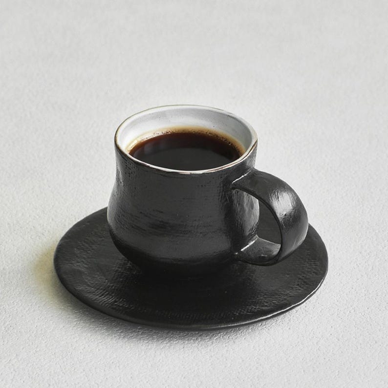 K&ouml;nnte beinhalten: Eine schwarze Keramik-Kaffeetasse mit passender Untertasse. Die Tasse ist mit dunklem Kaffee gef&uuml;llt und hat einen wei&szlig;en Rand. Tasse und Untertasse haben eine strukturierte Oberfl&auml;che, der Griff ist abgerundet.