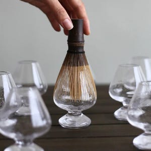 May include: A clear glass matcha whisk with a bamboo handle and whisking tines, held by a hand. Several clear, stemmed glasses surround the whisk, all on a dark wooden surface. The whisk is approximately 13 cm tall.
