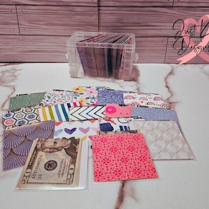 May include: A clear plastic storage box filled with colorful patterned paper squares. The box is sitting on a white marble surface. There are several more paper squares outside of the box, some of which are layered on top of each other. A $20 bill is visible in one of the squares.
