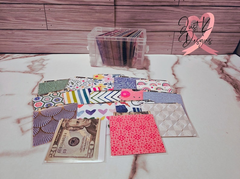 May include: A clear plastic storage box filled with colorful patterned paper squares. The box is sitting on a white marble surface. There are several more paper squares outside of the box, some of which are layered on top of each other. A $20 bill is visible in one of the squares.