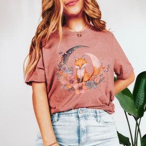 May include: A mauve t-shirt featuring a graphic of a fox sitting on a crescent moon surrounded by flowers. The fox is orange and white with a bushy tail.