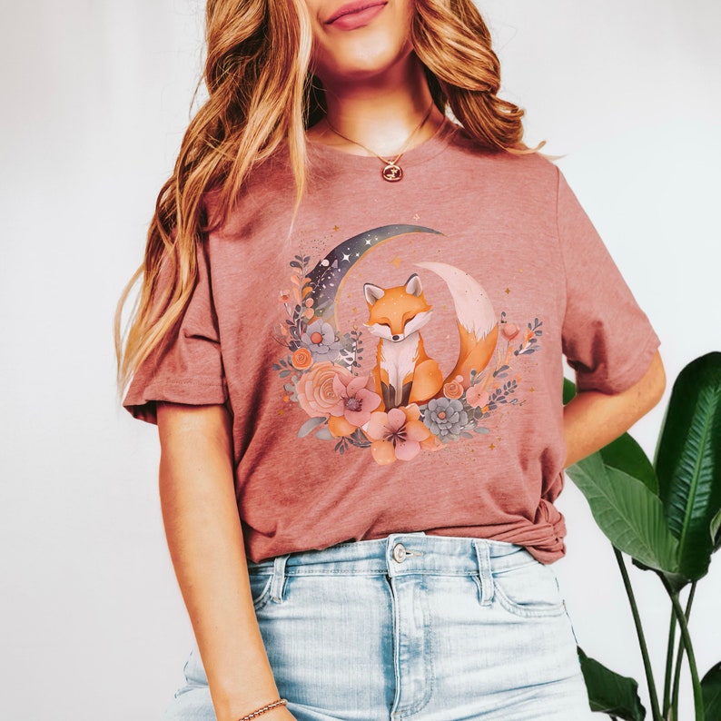 May include: A mauve t-shirt featuring a graphic of a fox sitting on a crescent moon surrounded by flowers. The fox is orange and white with a bushy tail.