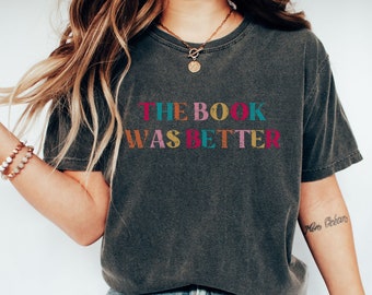 May include: Dark gray t-shirt with the text "THE BOOK WAS BETTER" in colorful letters. The shirt has a vintage, worn look. The text is in pink, teal, orange, yellow, and green.