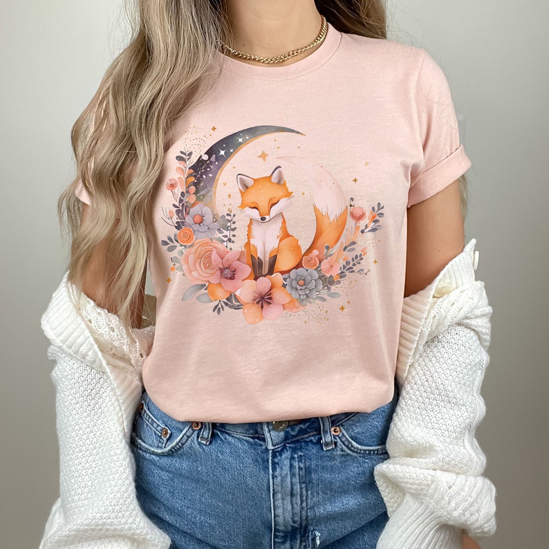 May include: A pink t-shirt featuring a graphic of a fox sitting on a crescent moon surrounded by flowers. The fox is orange and white with a bushy tail.