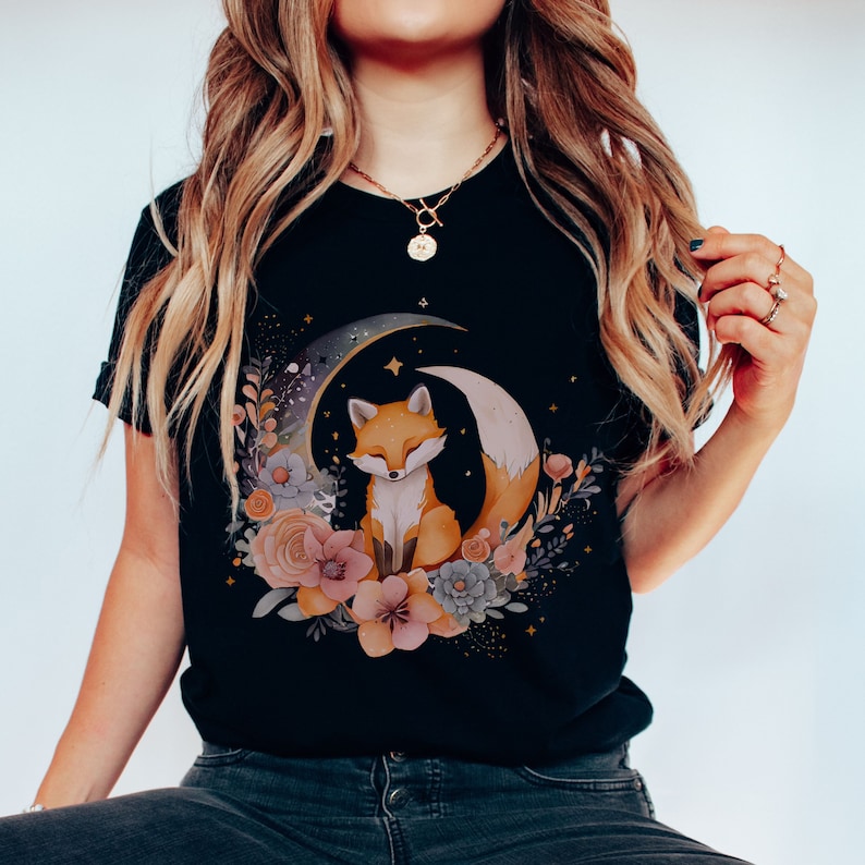 May include: Black t-shirt featuring a graphic of a fox sitting on a crescent moon surrounded by flowers. The fox is orange and white, and the flowers are pink, blue, and green.