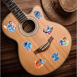 K&ouml;nnte beinhalten: Akustikgitarre mit mehreren bunten Aufklebern mit der Figur Stitch. Die Aufkleber zeigen Stitch in verschiedenen Posen. Ein brauner Cowboyhut liegt auf dem Korpus der Gitarre, vor einem Holzuntergrund.