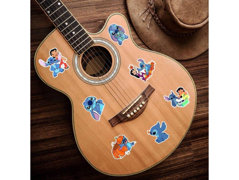 K&ouml;nnte beinhalten: Akustikgitarre mit mehreren bunten Aufklebern mit der Figur Stitch. Die Aufkleber zeigen Stitch in verschiedenen Posen. Ein brauner Cowboyhut liegt auf dem Korpus der Gitarre, vor einem Holzuntergrund.