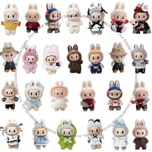 May include: A collection of small, plush bunny figures, each dressed in unique outfits. The figures feature bunny ears and are in various colors, including yellow, red, white, and blue. The text "Fun Doodle Studio" is visible.
