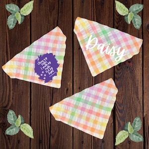 May include: Three pastel plaid dog bandanas. One bandana has the name "Daisy" in white script. Another bandana has the text "Clean my Path! I walk by Faith Not by Sight" in a purple circle with paw prints.