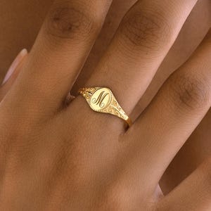 May include: A gold signet ring with an oval face and the letter "M" engraved in the center. The ring has a detailed floral design around the face and on the band. The ring is shown on a person's finger.