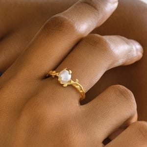 May include: A gold-colored ring featuring a central, round, white gemstone. The ring's band is adorned with delicate, leaf-like designs. The ring is shown on a person's finger.