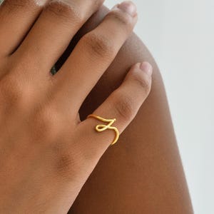 May include: A gold-colored ring in the shape of the letter "Z" is worn on a finger. The ring is made of a thin wire and has a minimalist design. The ring is on a hand with a light skin tone, with a green shirt in the background.