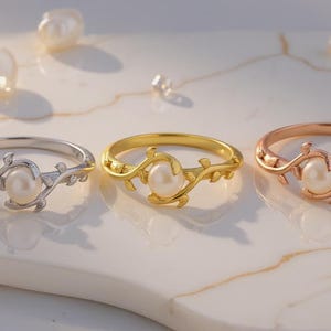 May include: Three rings with a pearl center and a delicate vine design. The rings are silver, gold, and rose gold. Scattered pearls and small crystals surround the rings on a white marble surface.