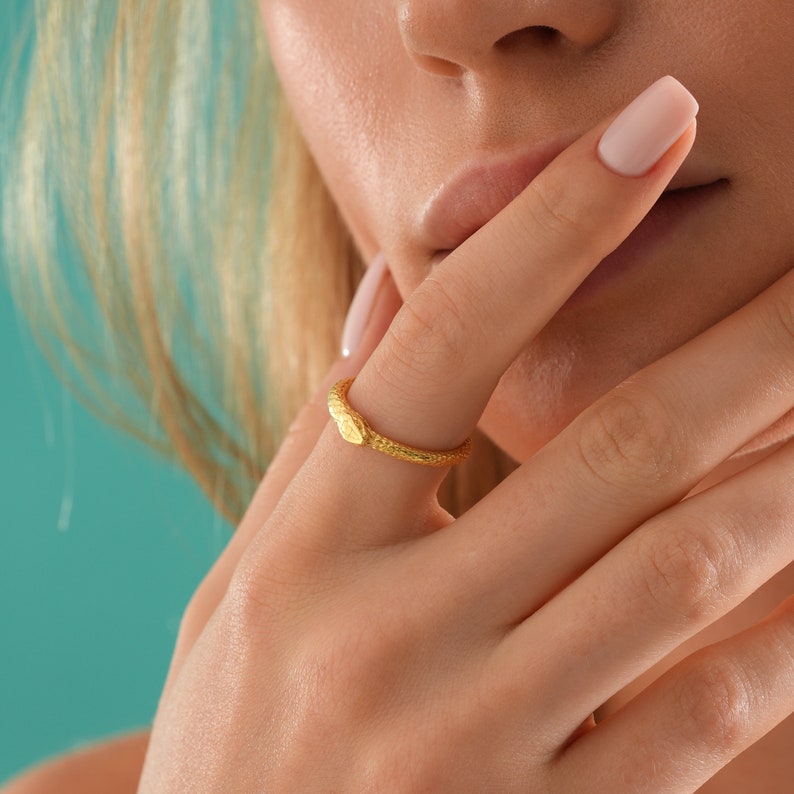May include: A gold snake ring with a textured surface. The ring is worn on the finger of a person with a light complexion.