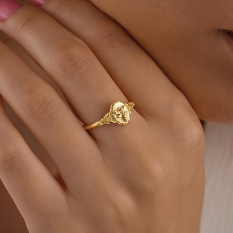 May include: A gold ring featuring a dragonfly design. The ring has an oval-shaped face with a detailed dragonfly and a delicate band with leaf accents. The ring is a warm gold color.
