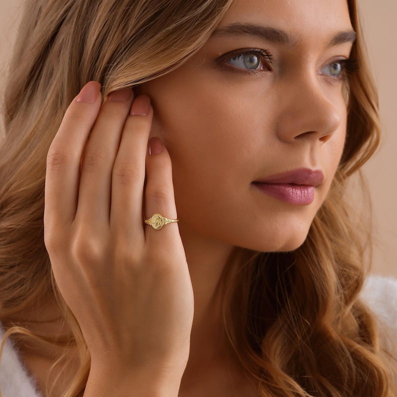 K&ouml;nnte beinhalten: Ein goldener Ring mit einem detaillierten, ovalf&ouml;rmigen Design. Der Ring befindet sich an einem Finger, und das Design scheint ein Blumen- oder abstraktes Muster zu sein. Der Ring ist der Fokus, mit der Hand und dem Gesicht der Person im Hintergrund.